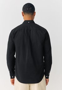 Črna majica z dolgimi rokavi, ostro ovratnico, zadnjo gubo in zapenjanjem na gumbe na manšetah. Gladka tekstura in rahlo ukrivljen spodnji rob.