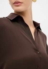 Camisa de seda marrón con cuello, frontal de botones y textura suave. Presenta un escote en V profundo y elegantes detalles de sastrería.