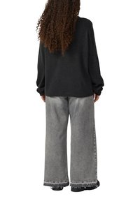 Persoon met lang krullend haar, gekleed in een losse zwarte trui en wijde, vervaagde grijze jeans met rafelige onderkanten, staand met de rug naar ons gekeerd.