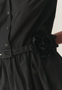 Čierne šaty s priliehavým telom, veľkým kvetinovým akcentom v páse, gombíkmi a z gatherovanou sukňou. Vyrobené z hladkej, ľahkej tkaniny.