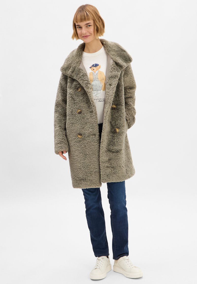 Taupe nepbontjas met brede kraag, dubbele knopen en twee zijzakken, gecombineerd met een grafisch shirt en spijkerbroek.