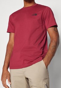 Camiseta de algodón color burdeos con cuello redondo y mangas cortas, con un pequeño logo negro en el pecho. Combinada con pantalones cortos de color claro.