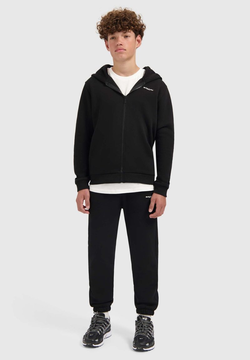 Zwarte ritshoodie met een wit logo, gecombineerd met zwarte sweatpants en zwarte sportschoenen. Gebalanceerde pasvorm met een capuchon en manchetten.