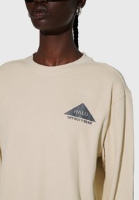 Ljusbeige långärmad skjorta med texturerat tyg, med en grå triangulär logotyp med texten "HALO OFF DUTY GEAR" på bröstet.