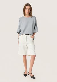 Grå kortærmet top med subtile udsmykninger, parret med hvide shorts til knæene. Sorte stroppede spidse flade sko fuldender looket.