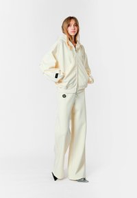 Chaqueta con capucha de color crema y cierre, con mangas amplias, combinada con pantalones de pierna ancha a juego. Presenta un delicado detalle de costuras y logotipos.