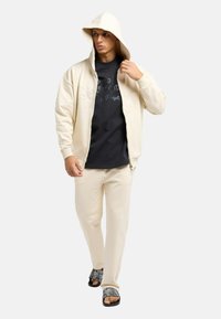 Cremefarvet hættezipper sweatshirt over en sort t-shirt, parret med cremefarvede joggers og mønstrede slidesandaler, med en afslappet pasform.