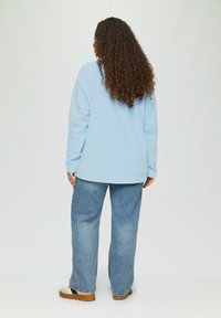 Lichtblauwe sweatshirt met lange mouwen, ontspannen pasvorm, gecombineerd met losvallende blauwe jeans en beige platform schoenen, gezien vanaf de achterkant.