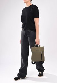 Mochila verde oliva con un diseño de roll-top, bolsillo frontal con cremallera y correas negras, sostenida por una persona que lleva una camiseta negra y vaqueros.