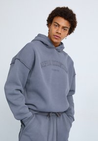 Szara bluza z kapturem i dopasowane dresy wykonane z miękkiego materiału, z logo na klatce piersiowej i sznurkiem w pasie. Luźny krój.