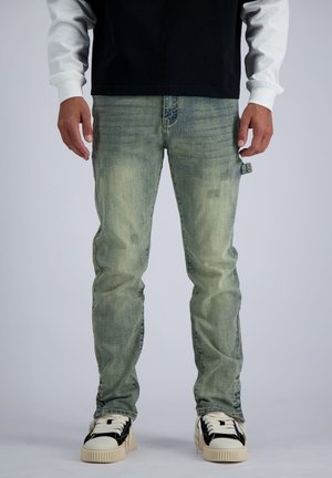 Mann trägt hellblaue ausgewaschene Jeans, schwarz-weiße Sneaker und ein schwarzes Shirt über einem weißen Langarmshirt, steht vor einfarbigem Hintergrund.