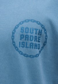 Μπλε ύφασμα με σκούρο μπλε κείμενο «Νότιο Νησί Πάντυ» μέσα σε σχεδιασμό κυκλικής αλυσίδας.