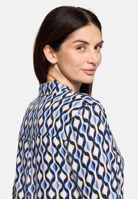 Chemise à manches longues présentant un motif de vagues bleu, beige et noir sur un tissu léger, avec un col et des plis à l'arrière.