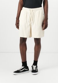 Cremefarvede shorts med et tekstureret, bølget mønster, der har en elastisk talje og snørebånd. Sammen med sorte sneakers.