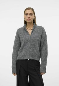 Grå glidelåsgenser med en strukturert strikkefinish, ribbestrikkede mansjetter og høy krage, kombinert med svarte plissert bukser.