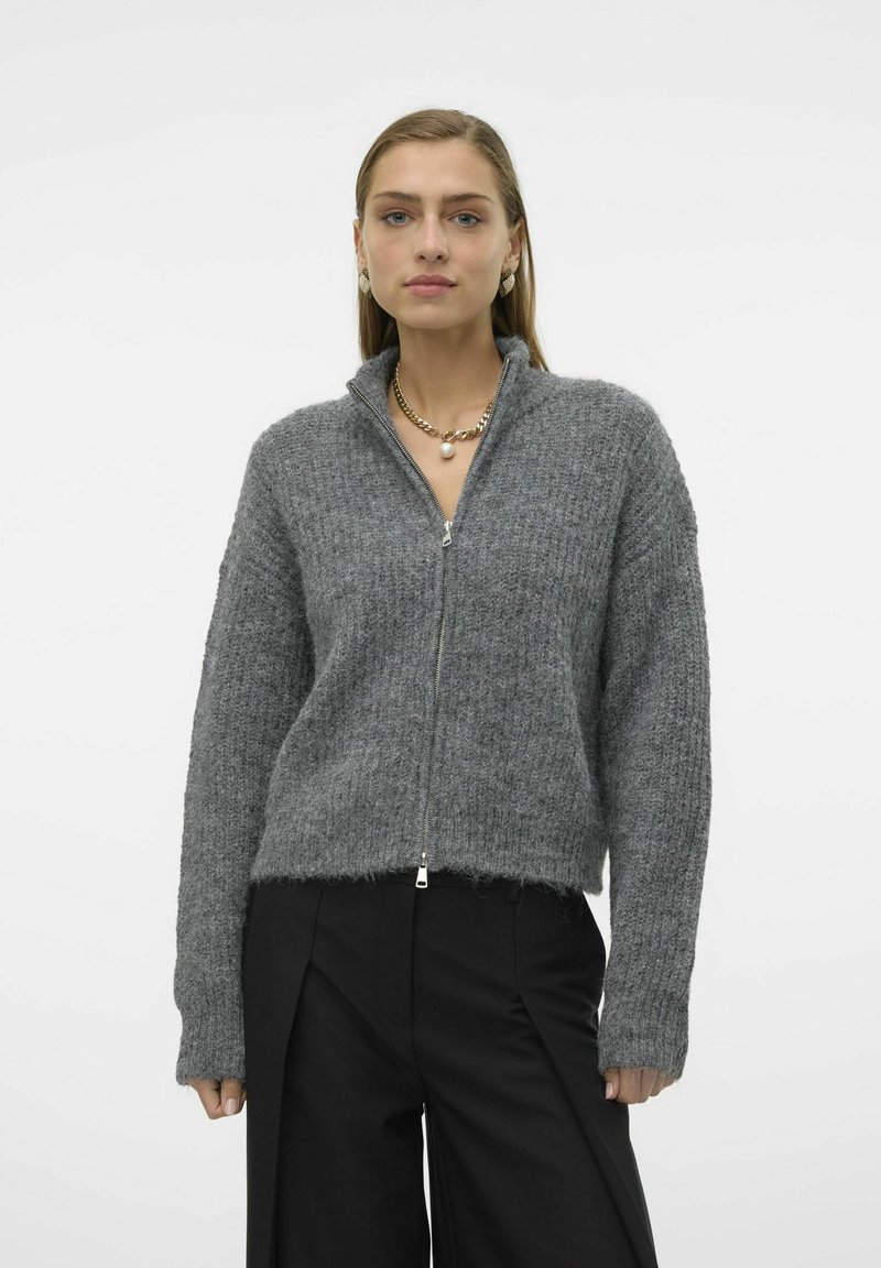 Grå glidelåsgenser med en strukturert strikkefinish, ribbestrikkede mansjetter og høy krage, kombinert med svarte plissert bukser.