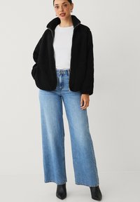 Sort fleecejakke med høj krave og forlommer, parret med lyseblå viddejeans og sorte spidse ankelstøvler.