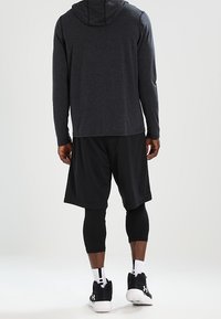 Czarna bluza z długim rękawem i kapturem, zestawiona z czarnymi siatkowanymi szortami oraz czarnymi legginsami kompresyjnymi. Obuwie stanowią czarne sportowe buty.