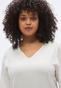 Λευκό τοπ με V-λαιμόκοψη και μακριά μανίκια, με υφή στην επιφάνεια. Το άτομο φορά λεπτό, μεταλλικό κολιέ και έχει σγουρά μαλλιά. Ουδέτερο φόντο.