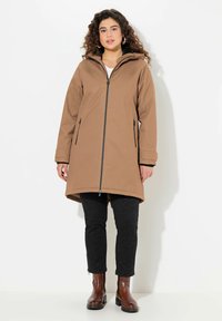 Manteau imperméable marron avec fermeture éclair sur le devant, capuche et deux poches latérales. Porté par-dessus un pantalon noir à motifs et des bottines marron. Tissu texturé.