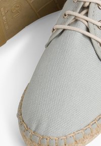 Scarpa in tela grigia chiara con cuciture beige, punta rotonda e design con lacci. Presenta una superficie testurizzata e una suola in fibra naturale.