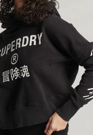 Person wearing a black hoodie with "SUPERDRY" and Japanese characters in white print, standing with one hand on their hip.