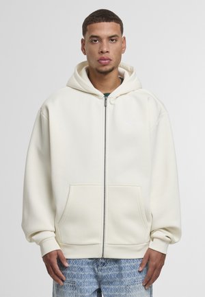 Cream zip-up hoodie in soft fabric with a large hood and kangaroo pockets. Features a subtle logo on the chest and ribbed cuffs.