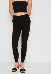 Černé upnuté kalhoty s hladkým povrchem, tapered nohavicemi a středně vysokým pasem. Spárovány s černými sandály na vysokém podpatku se šněrováním.