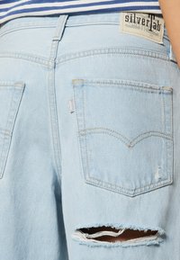 Lyseblå denim jeans med slidte detaljer, herunder et stort hul, to baglommer og et mærke på livet.