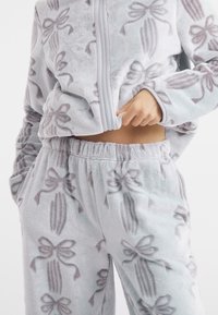 Lys blå loungewear sæt i fleecemateriale med diskrete sløjfemønstre, lynlåsjakke og bukser med elastisk talje for komfort.