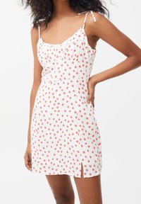 Frau in einem weißen ärmellosen Kleid mit rotem Punktmuster und Seitenschlitz, die mit einer Hand auf ihrer Hüfte posiert.