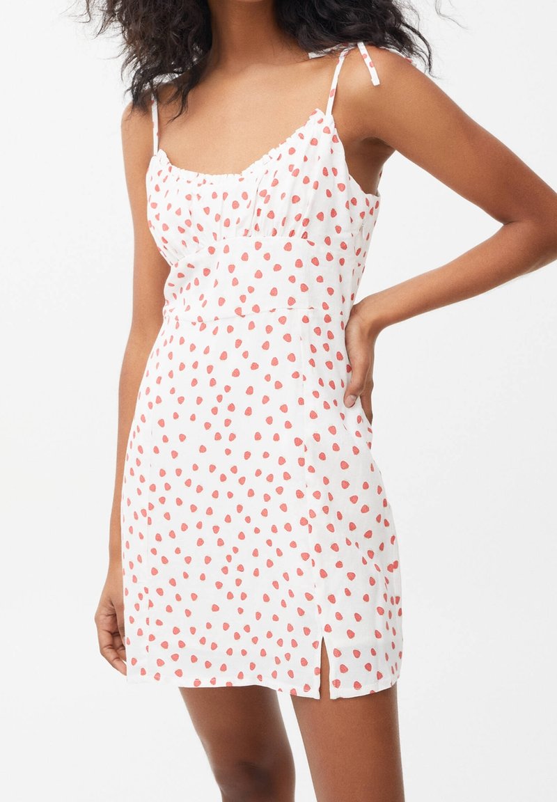 Femme portant une robe sans manches blanche avec un motif à pois rouges et une fente sur le côté, posant avec une main sur la hanche.