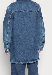 Long blue denim jacket with a relaxed fit, featuring visible stitching, button accents on cuffs, and a slightly longer back hem.