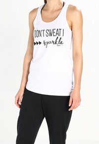 Débardeur blanc avec un texte noir indiquant "NE SUEZ PAS, JE BRILLE", doté d'un design dos nageur et d'une coupe décontractée ; porté avec un pantalon noir.