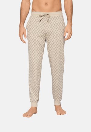 Mannelijke torso en benen, gekleed in beige loungebroek met patroon en trekkoord, met omslagen bij de enkels, staand op blote voeten tegen een effen achtergrond.