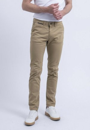 Homme portant un pantalon beige slim et des baskets blanches, debout devant un fond clair uni avec les mains près de la taille.