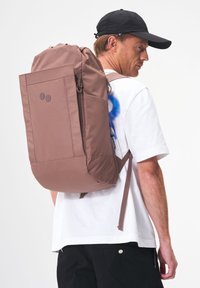 Brauner Stoffrucksack mit gepolsterten Schulterträgern, rechteckiger Form, Vordertasche und zwei runden Akzenten. Trägt eine Person in einem weißen Hemd.