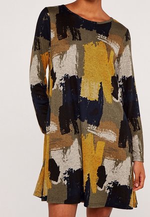 Femme portant une robe à manches longues et ample avec des motifs abstraits de coups de pinceau en jaune, noir, marron, gris et bleu marine.