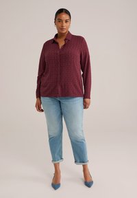 Camisa de manga larga color burdeos con un patrón sutil, botones en la parte delantera y un ajuste holgado, combinada con jeans azul claros arremangados y tacones azules.