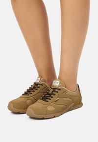 Zapatillas de malla y cuero ante marrón con cordones negros, suelas de goma marrón claro y logotipo en la lengüeta. Diseño bajo para uso casual.