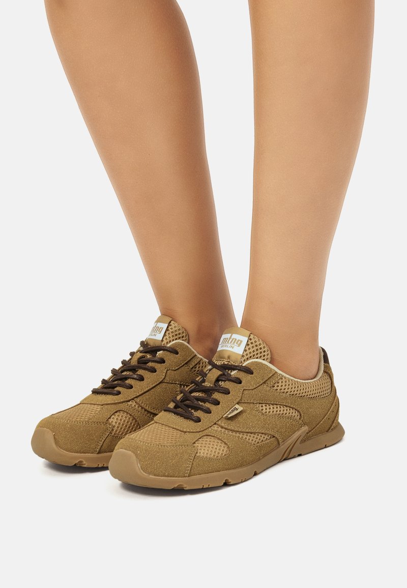 Zapatillas de malla y cuero ante marrón con cordones negros, suelas de goma marrón claro y logotipo en la lengüeta. Diseño bajo para uso casual.