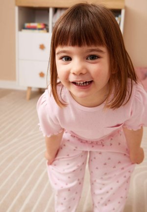 Uśmiechnięta młoda dziewczyna o brązowych włosach, ubrana w różową piżamę, lekko pochylona do przodu w delikatnie oświetlonym pokoju z wzorzystym dywanem i białymi meblami.