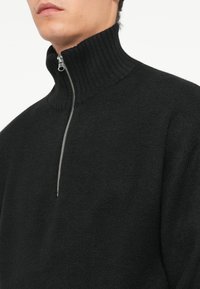 Sweat à zip noir avec un col côtelé haut, fabriqué à partir d'un matériau tricoté texturé. Doté d'une fermeture éclair argentée au niveau du cou.
