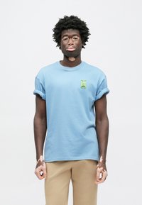 Gaiši zils kokvilnas T-krekls ar uzlocītām piedurknēm un mazu zaļu karikatūras uzlīmi uz krūtīm, valkāts ar bēšām biksēm.