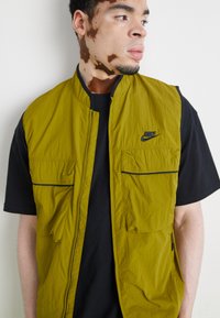 Nike alyvuogių žalio atspalvio liemenė su lengvu tekstūru, su keliais priekiniais kišenių, užtrauktuku ir juodu logotipu. Nešiojama virš juodo marškinėlių.