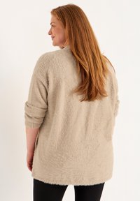 Beige, tekstuuri-pullover med et afslappet fit, trekvart ærmer og en rund nederste kant, båret over sorte leggings.