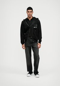 Sweatshirt preta com fecho na frente e bolso, combinada com jeans pretos de perna reta e ténis pretos. Design simples e casual, sem padrões.