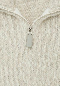 Kremasto bež pulover iz pletenega materiala z zadrgo na ovratniku, grobo teksturo in sivim usnjenim potegom zadrge z vtisnjenim logotipom.