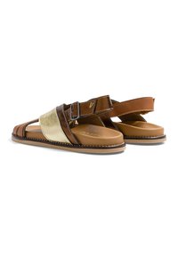 Paire de sandales en cuir marron avec des brides dorées, des boucles latérales réglables et des semelles plates beiges sur fond blanc.