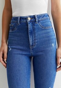 Mujer vistiendo jeans de mezclilla azules de tiro alto con un ligero desgaste y una camiseta blanca de punto sin mangas, con las manos relajadas a los lados.